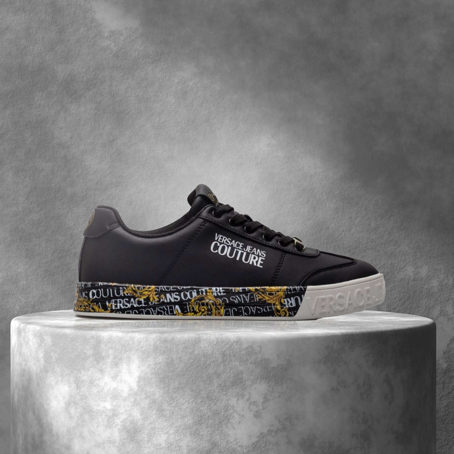 Black Versace Jeans Couture sneaker with logo and gold detailing on a concrete pedestal, side view.